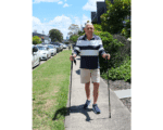 Older man walking with two trekking poles on a sunny sidewalk, smiling, wearing a striped polo shirt and beige shorts.