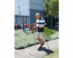 Older man walking with trekking poles on a sidewalk beside a landscaped area and a fence with red fire hydrant valves behind him.
