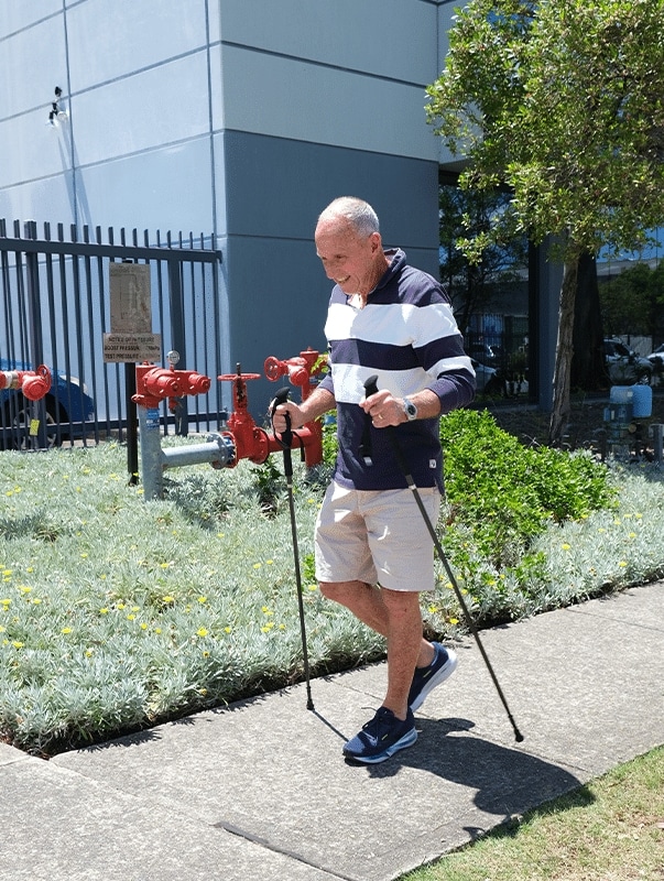 Older man walking with trekking poles beside a building and red utility valves, enjoying a sunny day